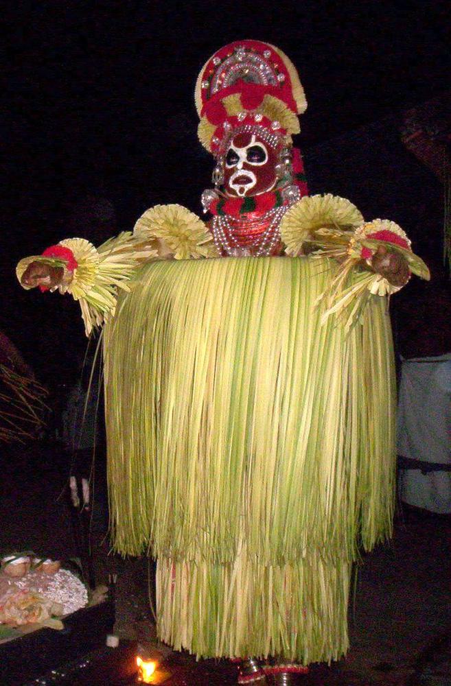 A Theyyam performer at night in towering costume and grass skirt